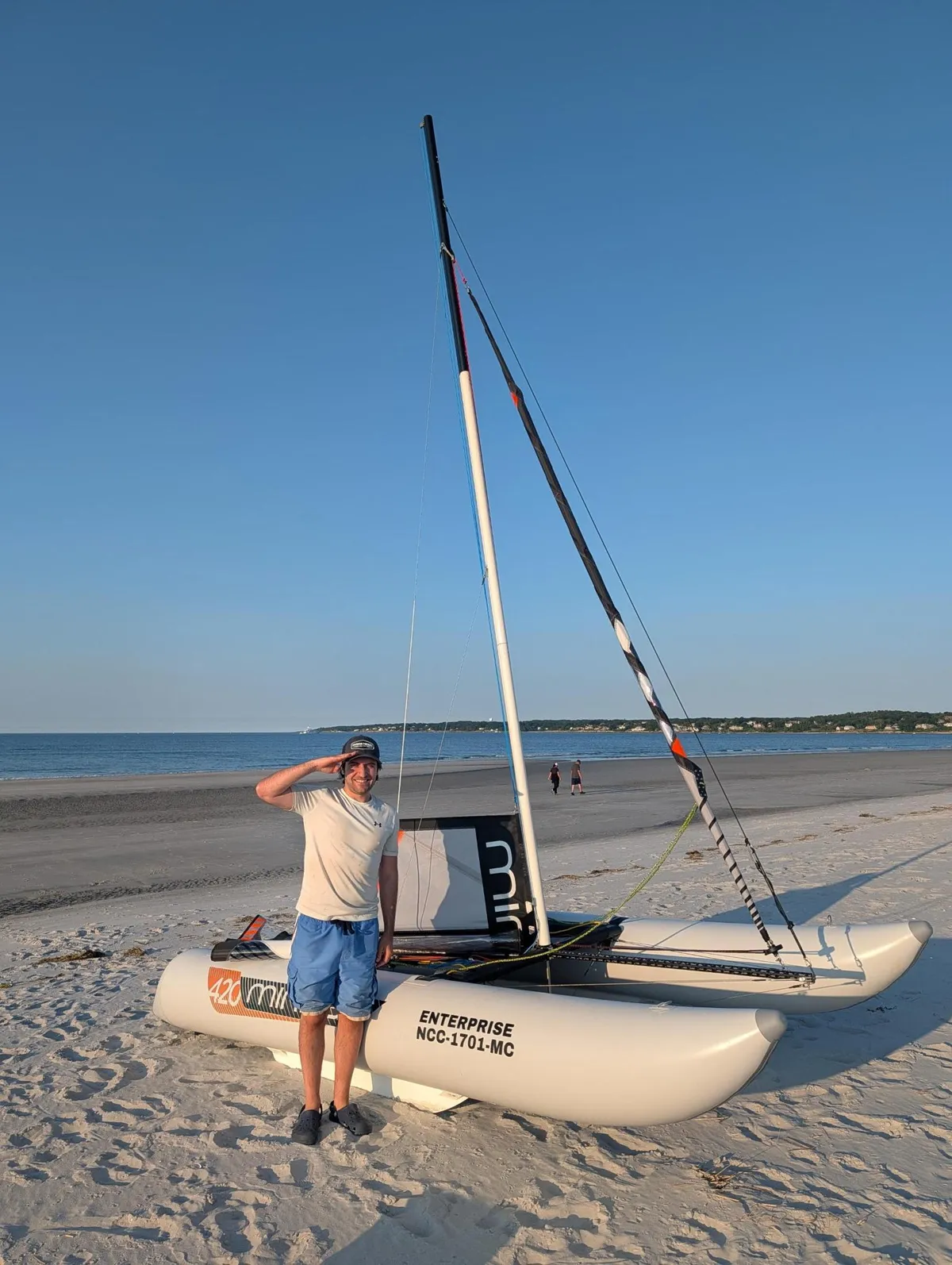 Robert Grassi standing on a beach beside a small beached catamaran sailboat, with the sea behind him.
